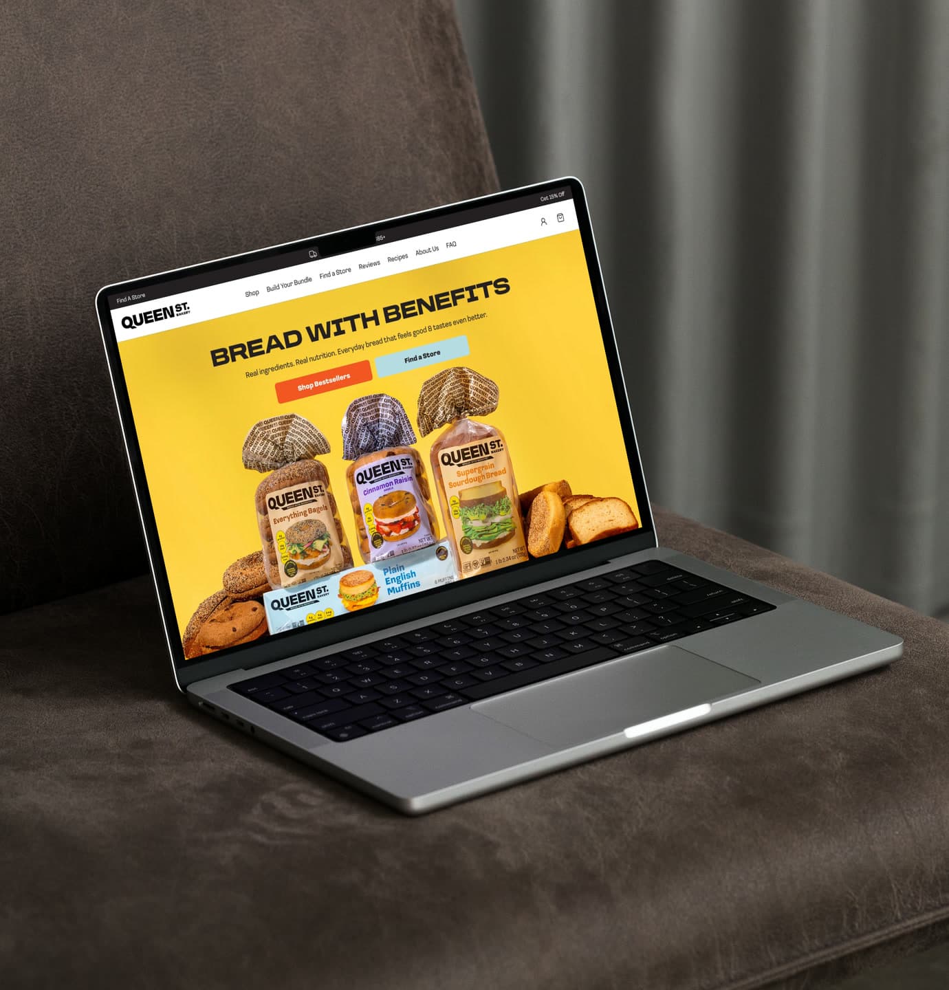 The Queen Street Bakery homepage open on a silver laptop resting on a brown leather couch, its yellow hero reading “Bread With Benefits” above three packaged loaves.